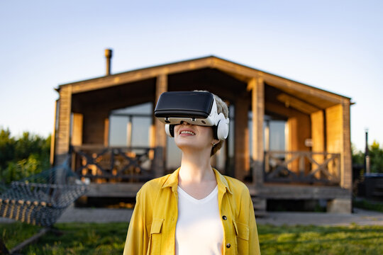 A young caucasian woman wearing a virtual reality headset and headphones enjoys an immersive experience in a natural outdoor setting near a wooden house