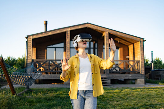 A caucasian woman wearing a virtual reality headset and headphones enjoys an immersive experience in a grassy outdoor setting near a wooden cabin