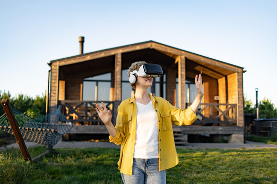 A young caucasian woman wearing a virtual reality headset and headphones enjoys an immersive experience in a natural outdoor setting near a wooden cabin