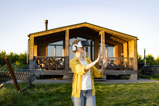 A young woman wearing a virtual reality headset and headphones is actively engaged in a virtual experience while standing in a grassy yard in front of a wooden house