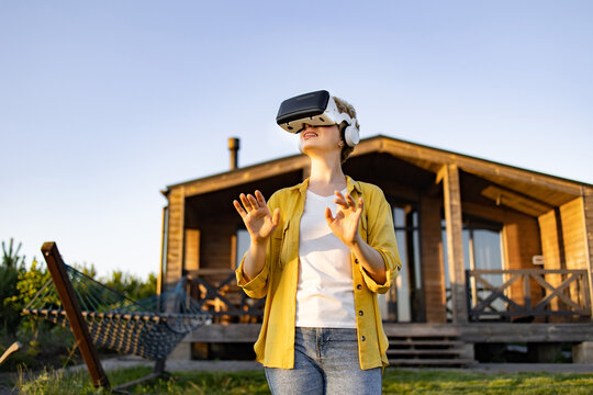 A young caucasian woman wearing a virtual reality headset and headphones enjoys an immersive experience in a natural outdoor setting near a wooden house