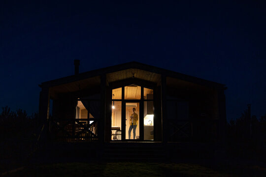A solitary caucasian woman stands inside a brightly lit modern cabin at night, looking out into the darkness