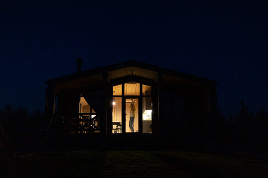 A young caucasian woman joyfully dances inside a brightly lit modern cabin at night, silhouetted against the dark sky