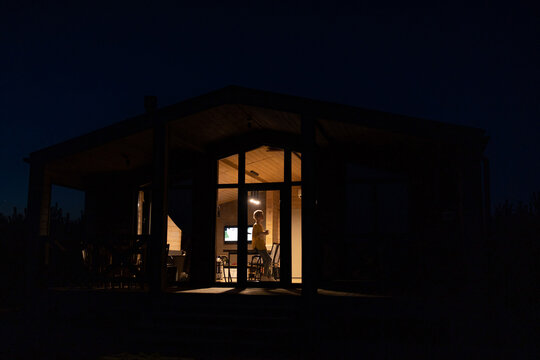 A solitary caucasian woman stands inside a modern cabin illuminated by warm interior lights against the dark night sky