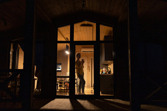A caucasian woman stands inside a cozy cabin, holding a warm mug and looking out a large window into the darkness