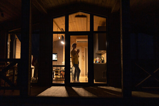 A caucasian woman stands inside a modern cabin, holding a mug and looking out a large window at night