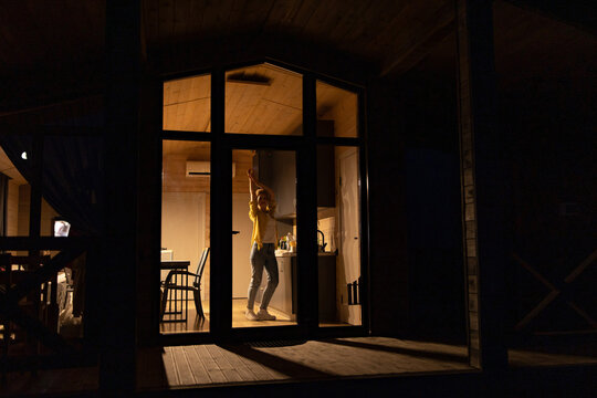 A happy young caucasian woman dances joyfully inside a modern cabin kitchen at night, illuminated by warm interior lights