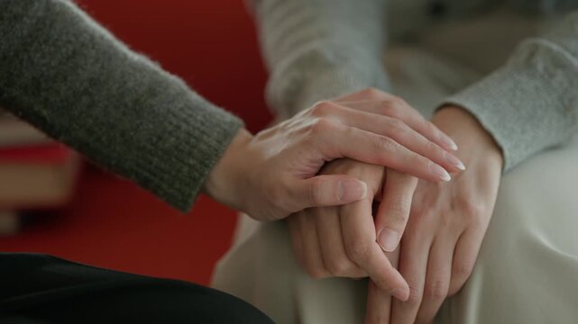 A close-up of two people's hands clasped together in a comforting or supportive gesture against a warm blurred background.