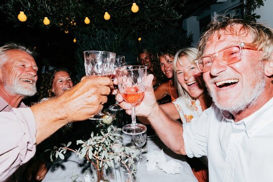 Cheerful senior friends toasting with wine glasses at a candid garden dinner party, featuring glowing string lights and a convivial mood for lifestyle and retirement imagery