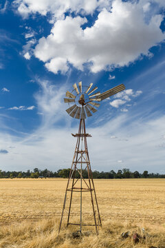 Aerial view of puffy white clouds in a blue sky above a farm windmill in the middle of a dry paddock