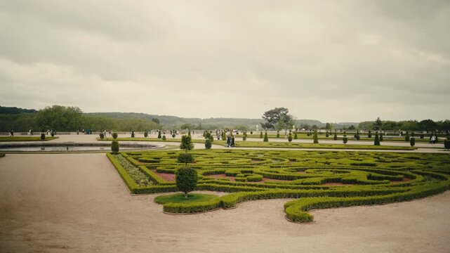 Sweeping aerial view of a grand formal garden featuring intricate parterre hedging, ornamental ponds, and manicured grounds with visitors exploring the landscape. Overcast European palace garden setti