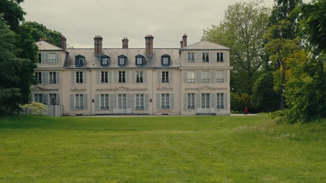 Wide-angle video of a grand neoclassical estate with cream-colored facade, mansard roof, and multiple chimneys, set against mature landscaping and verdant grounds. A solitary figure in red walks acros