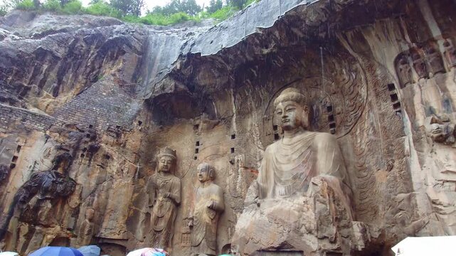 A sweeping wide-angle panoramic panning shot revealing the colossal rock-cut Vairocana Buddha statue and attendant figures carved into the limestone cliffside.