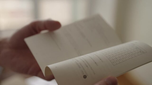 Close-up of hands flipping through Japanese manual and pointing to checklist grid on page, suggesting focused studying, reviewing instructions and reference material. Shooting in slow motion