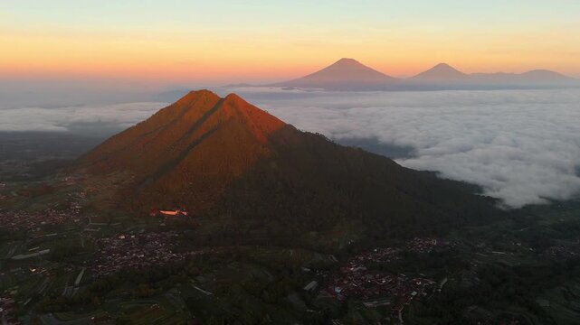 Aerial view of Mount Andong illuminated by golden sunrise light above a sea of clouds with lush volcanic slopes and village houses in Ngablak, Central Java, Indonesia.