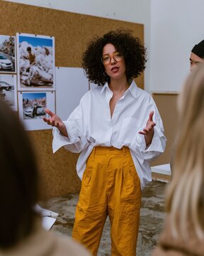 Focused Black woman in a white shirt and yellow trousers leading a creative team critique with expressive gestures in a bright design  for corporate storytelling