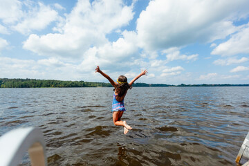 Obraz premium Girl jumps into a lake from a wooden dock on a sunny summer day. Child in swimsuit full of energy and excitement splashing into the water. Kid enjoys thrilling outdoor recreation and refreshing swim