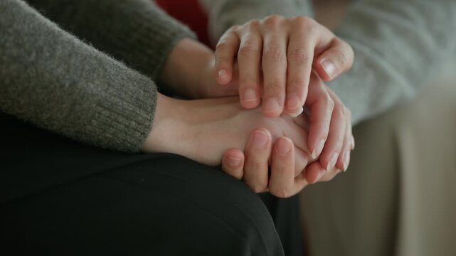 A close-up of two people's hands clasped together in a supportive gesture with a calm neutral background in an indoor setting.