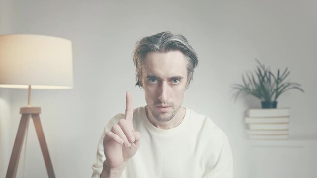 Serious adult man indoors raising index finger in denial gesture, seated against minimalist white background with tripod floor lamp, plant, and books
