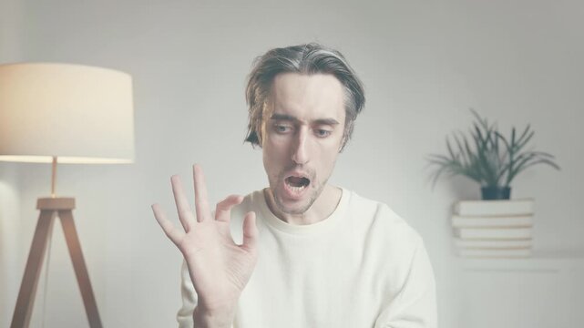 Confident adult man speaking to camera with OK hand sign in bright modern indoor space, minimalist decor with lamp and plants, suitable for lifestyle and communication themes