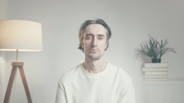 Young man sitting indoors in casual sweater, looking down with slight frown in minimalist softly lit interior with warm floor lamp and potted plant.