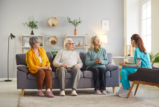 Group of elderly people sitting on sofa in nursing home and talking with friendly young nurse or psychologist woman during psychological therapy session. Psychotherapy for senior people.