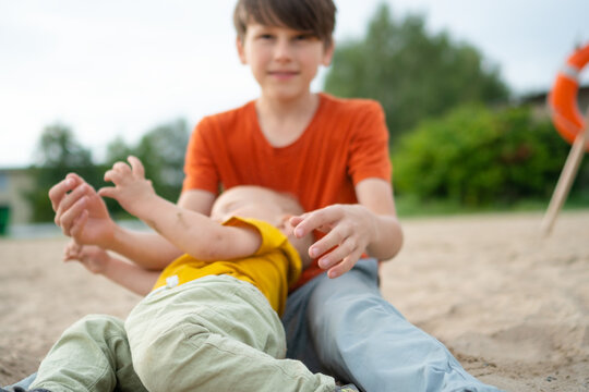Boy and toddler play on a beach together on a warm summer day. Child lying on back with older brother sitting nearby in the sand. Family shares joyful outdoor bonding and cheerful beach recreation
