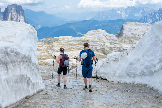 Rear view of two hikers with backpacks and trekking poles walking down a gravel path between high snow walls, couple enjoying active summer alpine adventure concept