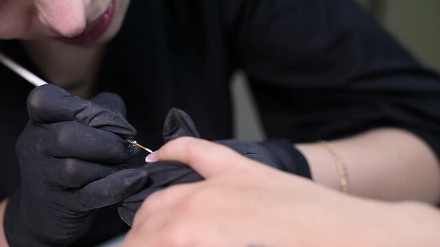 A professional manicurist wearing black gloves carefully paints a classic French manicure smile line. The close-up view highlights the precise brush technique on a client's nail.