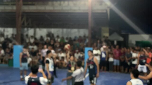 The referee is beginning a jump ball for basketball teams in a local league on an indoor, covered court. High angle, blurs.