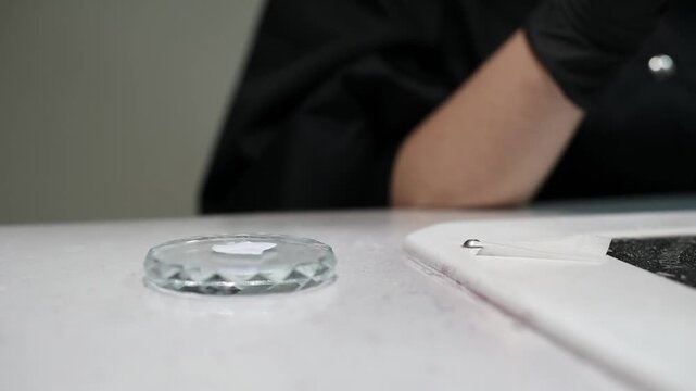 A close up shot of a small glass manicure dappen dish on a white table. A manicurist wearing black gloves prepares the workspace for a nail art procedure.