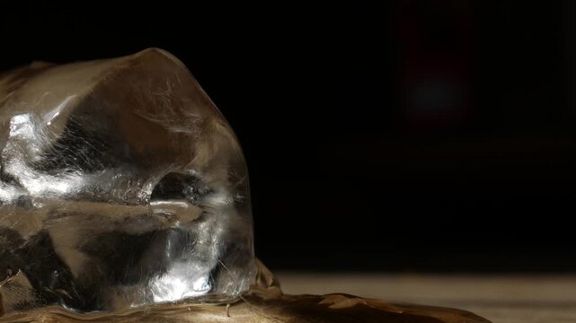 Single melting ice cube on rustic wooden table surface with dramatic sunlight shadow in dark moody still life macro photography