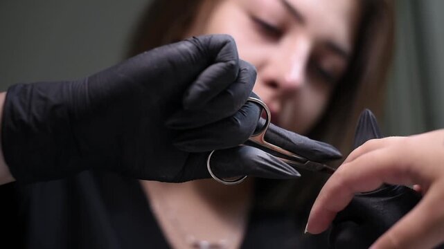 A professional nail technician in black gloves trims a client's cuticle with specialized scissors. The low angle close-up captures the precise hand care workflow.