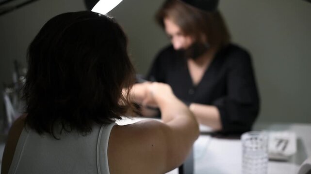 A professional manicurist carefully shapes and files a client's fingernails using a manual nail file. The over-the-shoulder medium shot shows the service in a salon setting.