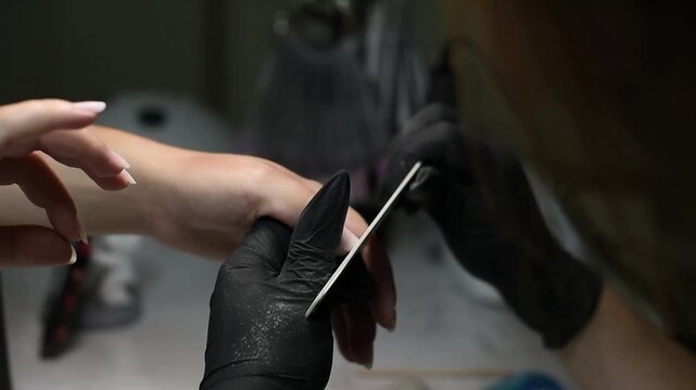 A professional manicurist wearing black gloves uses an emery board nail file to shape a client's fingernails. The close-up shot captures the precise filing technique in a salon.