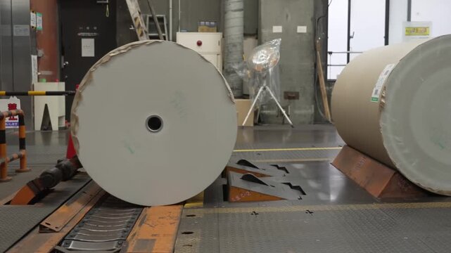Massive newsprint rolls resting on industrial rail conveyor inside large printing plant warehouse with red safety cone on factory floor