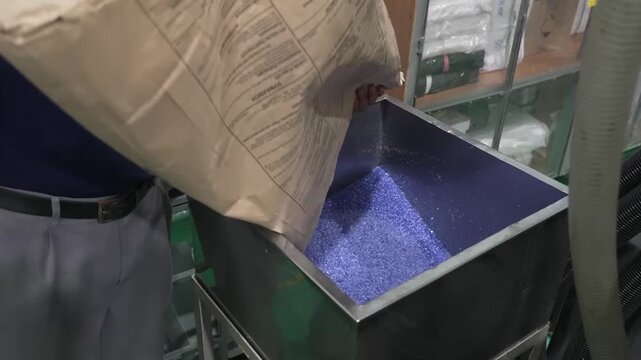 Factory worker pouring blue purple granular ink pigment from kraft bag into stainless steel mixing hopper inside ink manufacturing plant