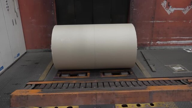 Single large newsprint roll with barcode inventory label resting on industrial rail conveyor inside paper mill ready for press production