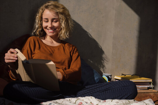 Portrait od smiling young woman sitting on bed reading a book