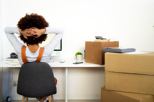 Afro woman with hand behind head relaxing by cardboard boxes in new home
