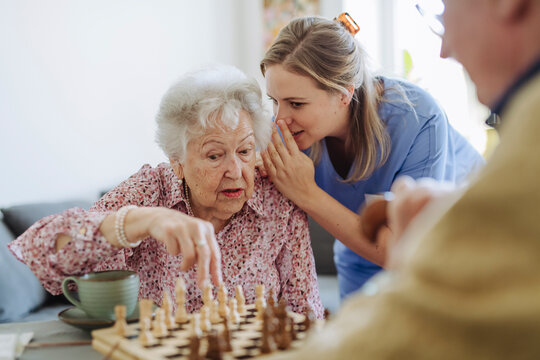 Healthcare worker whispering in senior woman's ear and playing chess at home