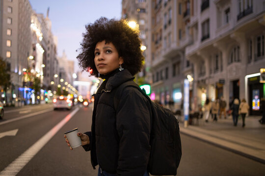 Young woman with disposable coffee cup crossing road while looking around in city at night