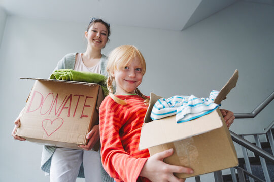 Smiling sisters with donation clothes in boxes moving downstairs