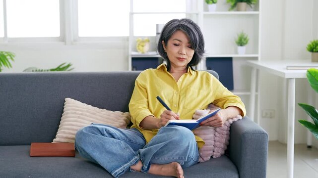 asian woman 50s years old write down in notebook diary stay at home flat rest relax spend free spare time in living room .