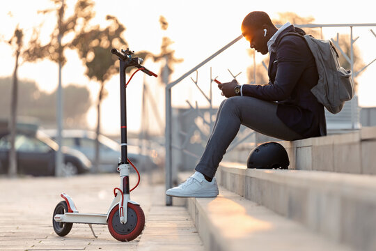 Young businessman with push scooter sitting on stairs at sunset using mobile phone and earphones