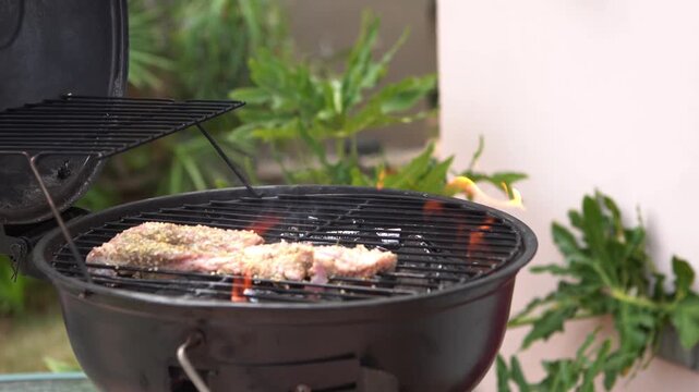 Close-up of seasoned meat being placed on a charcoal BBQ grill with tongs and open flames. Ideal for cookout, food prep, and outdoor cooking projects.