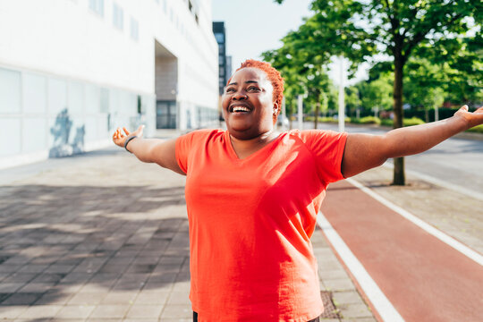 Smiling woman standing with arms outstretched on footpath