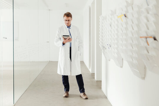 Mature man in lab coat standing in hallway looking at digital tablet