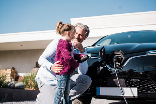 Happy girl holding electric plug standing by father in front of car on sunny day