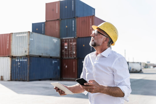 Businessman at cargo harbour, wearing safety helmet, using smartphone and digital tablet